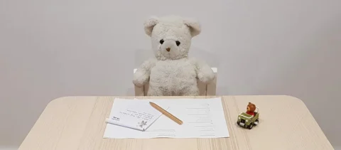 Teddy bear sitting at desk with papers and toy.